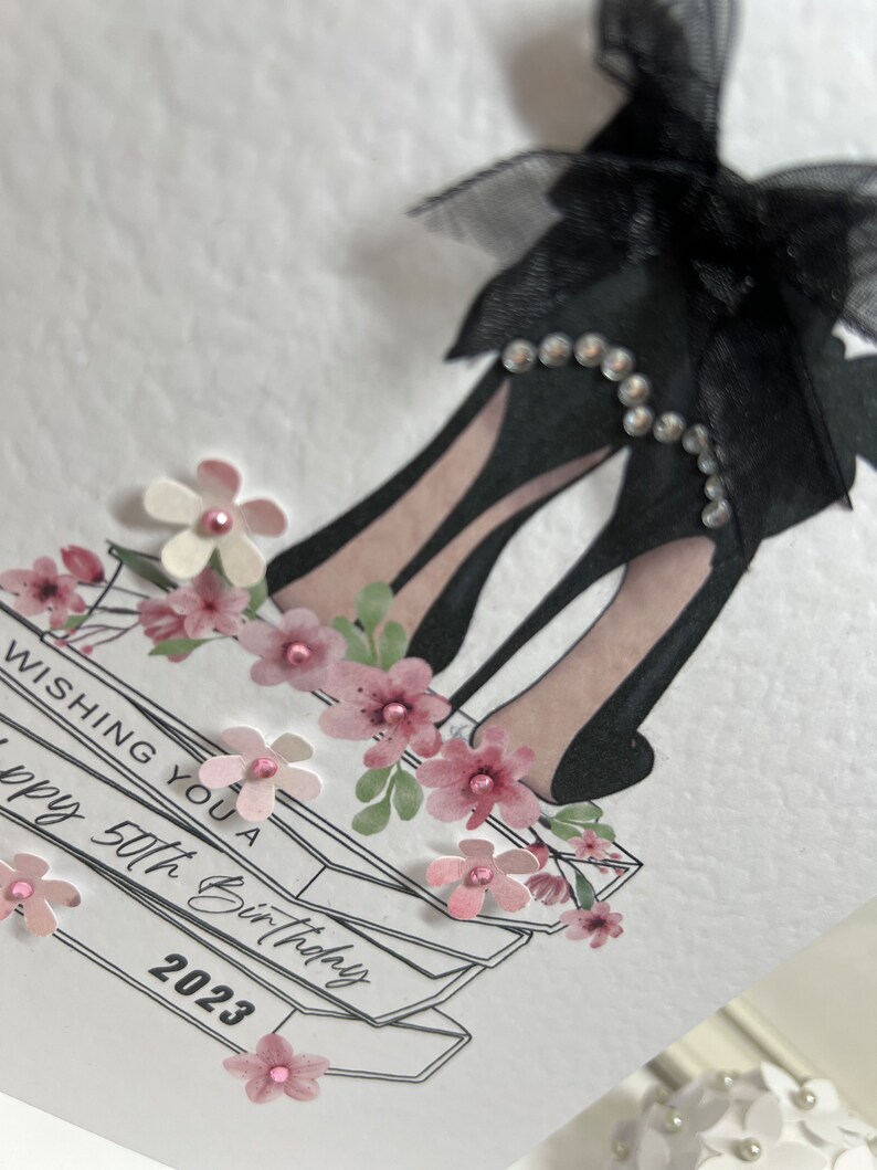 May include: A white card with a black high heel shoe with a black bow and rhinestones. The card has a stack of books with pink flowers and the text "Wishing you a happy 50th Birthday 2023".