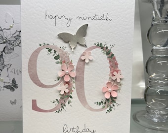 May include: A white card with the words "happy ninetieth birthday" written in black cursive. The number 90 is in pink with a floral design around it. A silver butterfly is on top of the card.