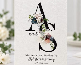 May include: A white wedding card featuring large black serif letters A and S connected by the word and. The letters are decorated with white, pink, and dark blue flowers, green leaves, and a white sheer ribbon. Text reads With love on your Wedding Day, Alistair & Stacey, 28th July 2025.