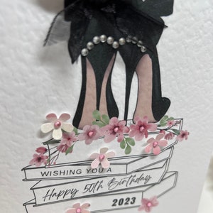 May include: A white card with a black bow and high heels on top of a stack of books. The card says "Wishing you a Happy 50th Birthday 2023".