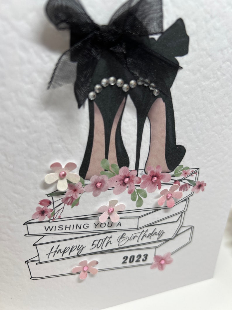 May include: A white card with a black bow and high heels on top of a stack of books. The card says "Wishing you a Happy 50th Birthday 2023".