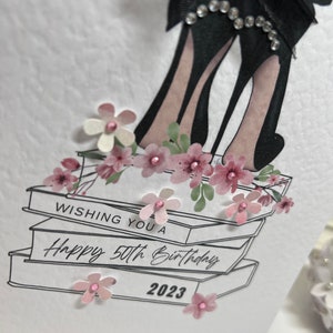 May include: A white card with a black and white illustration of high heels on top of books. The card has the text "Wishing you a Happy 50th Birthday 2023" and is decorated with pink flowers.