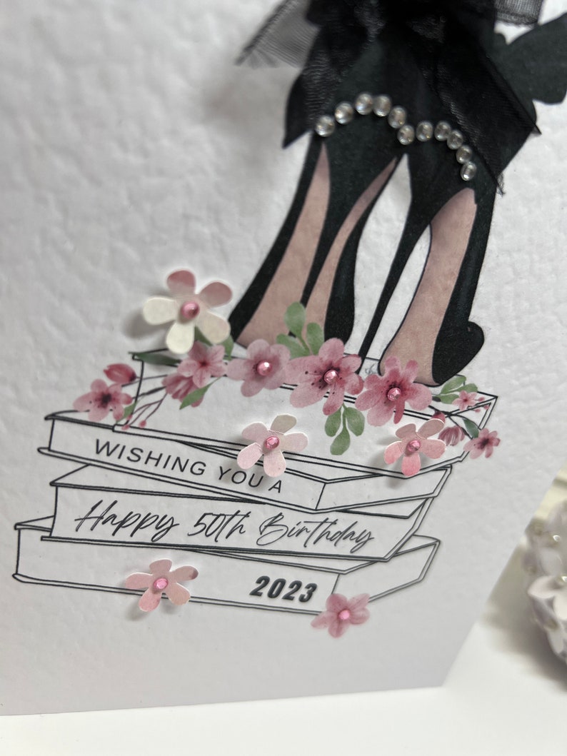 May include: A white card with a black and white illustration of high heels on top of books. The card has the text "Wishing you a Happy 50th Birthday 2023" and is decorated with pink flowers.