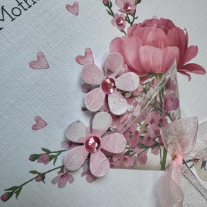 以下が含まれることがあります： 手作りの母の日カード。ピンクの紙の花、宝石の中心、大きなピンクの牡丹、小さなピンクのハートが白いテクスチャの背景に配置されています。「Mother's Day」の文字が黒で印刷されています。
