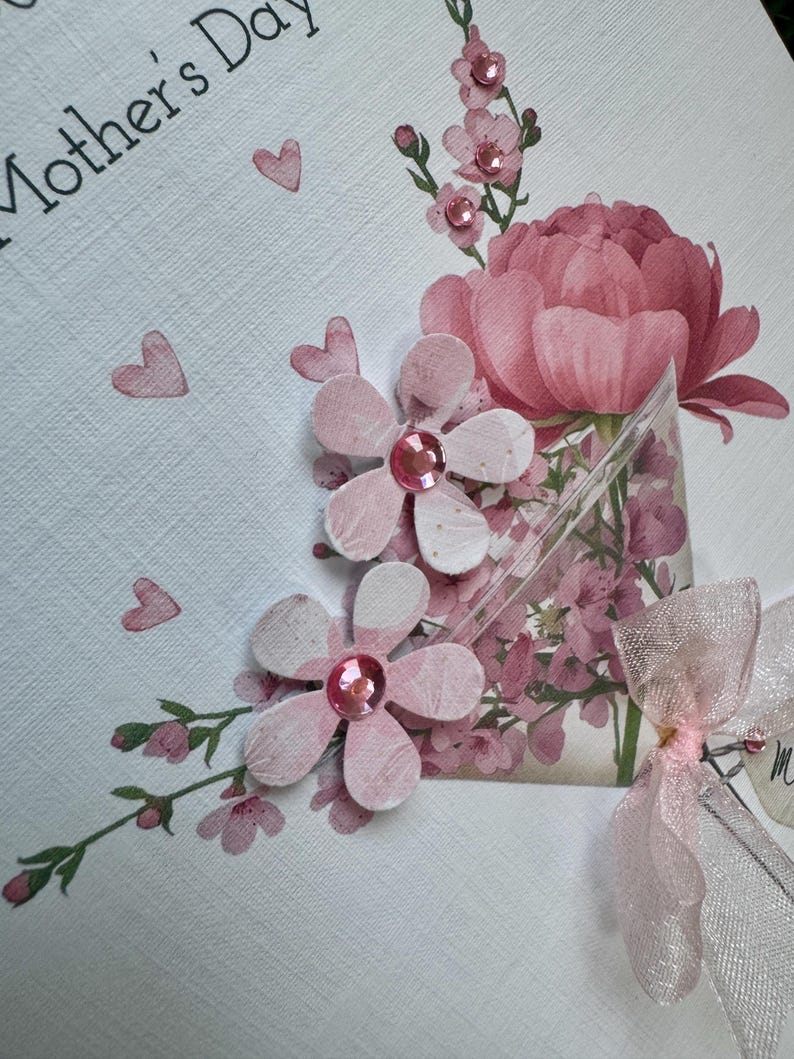 以下が含まれることがあります： 手作りの母の日カード。ピンクの紙の花、宝石の中心、大きなピンクの牡丹、小さなピンクのハートが白いテクスチャの背景に配置されています。「Mother's Day」の文字が黒で印刷されています。