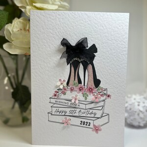 May include: A white card with a black and white illustration of a pair of high heels with a black bow on top. The heels are sitting on top of a stack of books. The card says "Wishing you a Happy 50th Birthday 2023".