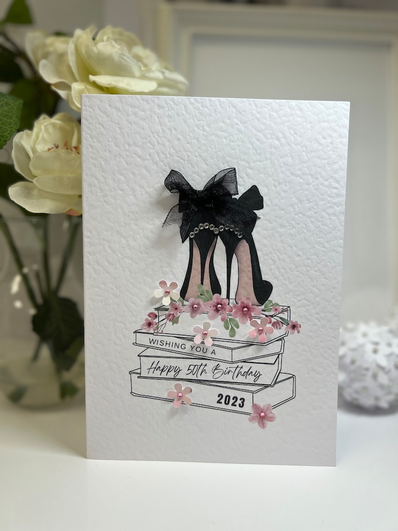 May include: A white card with a black and white illustration of a pair of high heels with a black bow on top. The heels are sitting on top of a stack of books. The card says "Wishing you a Happy 50th Birthday 2023".