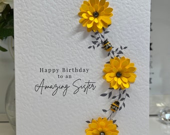 May include: A white card with a gray floral design and yellow flowers. The card says "Happy Birthday to an Amazing Sister".