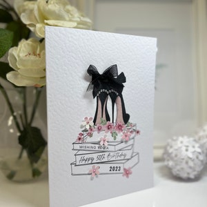 May include: A white card with a black and white illustration of a pair of high heels with a bow on top. The heels are on top of a stack of books with pink flowers. The card says "Wishing you a Happy 50th Birthday 2023".