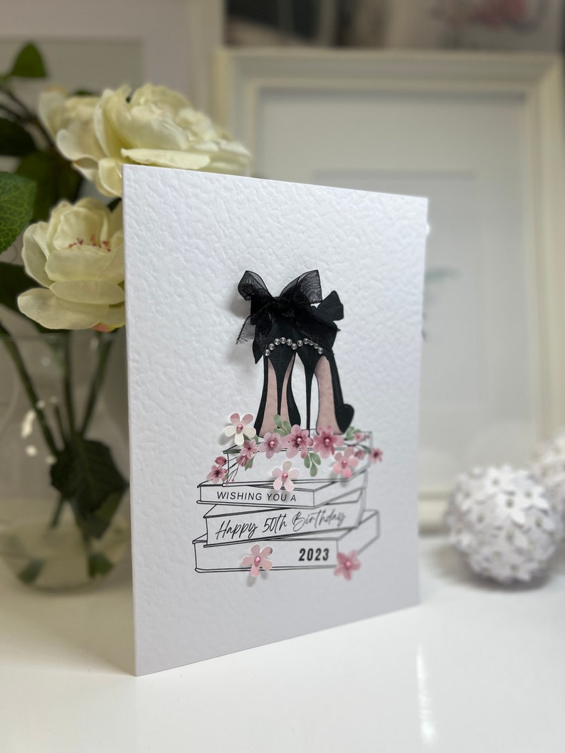 May include: A white card with a black and white illustration of a pair of high heels with a bow on top. The heels are on top of a stack of books with pink flowers. The card says "Wishing you a Happy 50th Birthday 2023".