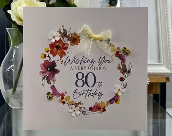 May include: A white card with a watercolor wreath of flowers and bees. The card says "Wishing you a very happy 80th birthday".
