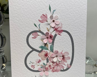 May include: A white card with a gray number 80 decorated with pink flowers and the text "With love on your 80th Birthday".