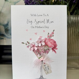 以下が含まれることがあります： 「With Love To A Very Special Mum」のメッセージが書かれた母の日カード。ピンクの花の装飾がマティーニグラスに入っており、「Happy Mother's Day 2028」と書かれたタグが付いています。カードはテクスチャ加工された白い背景です。