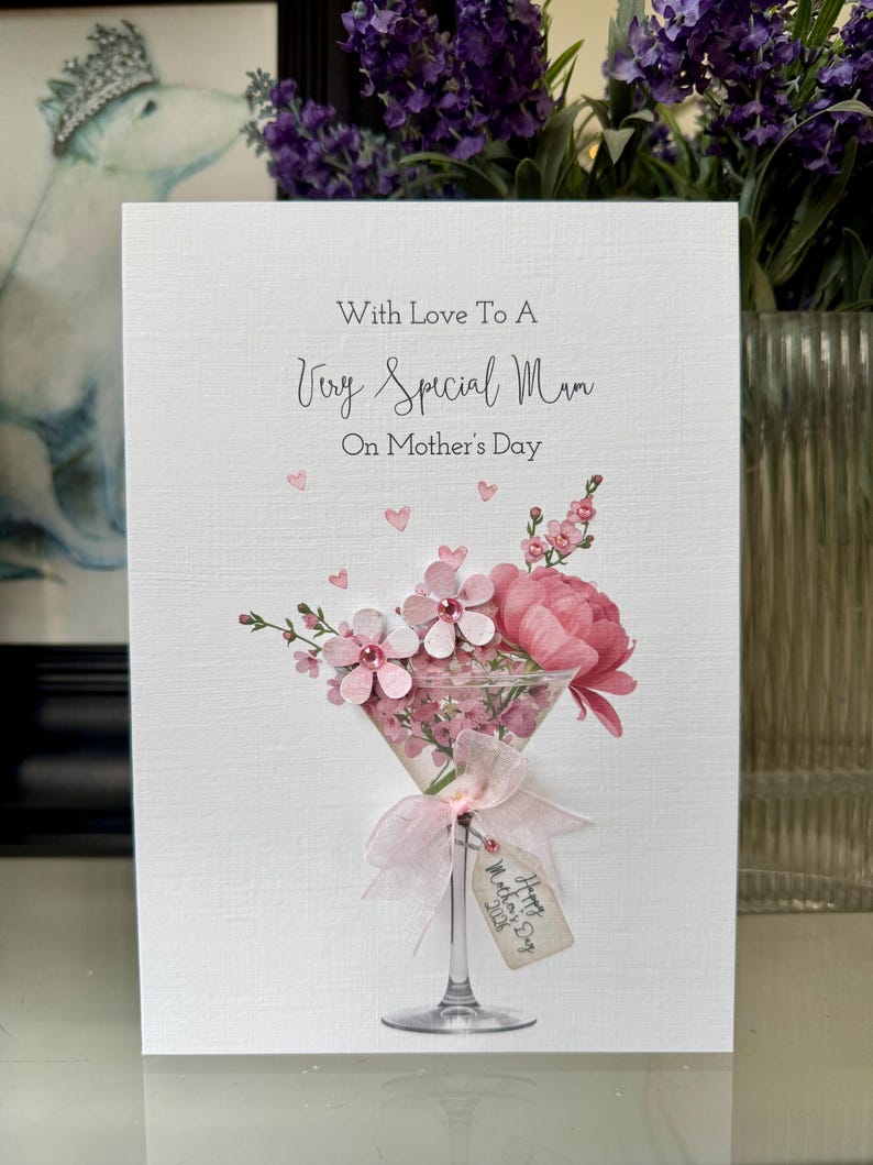 以下が含まれることがあります： 「With Love To A Very Special Mum」のメッセージが書かれた母の日カード。ピンクの花の装飾がマティーニグラスに入っており、「Happy Mother's Day 2028」と書かれたタグが付いています。カードはテクスチャ加工された白い背景です。