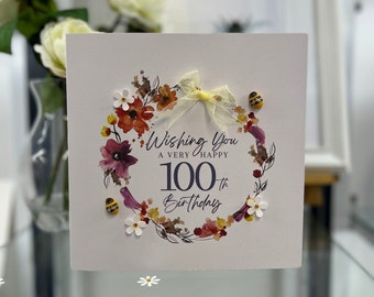 May include: A white card with a watercolor wreath of flowers and leaves in shades of red, orange, yellow, and purple. The wreath surrounds the number 100 with the word "th" in superscript. The text "Wishing You A Very Happy 100th Birthday" is written in black lettering. There are three small yellow and black bee figurines attached to the card.