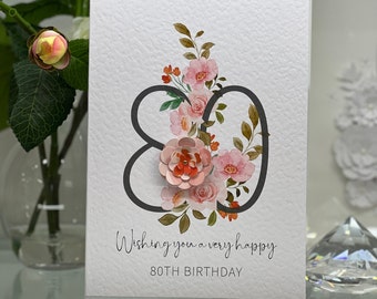 May include: A white card with a black number 80 decorated with pink and orange watercolor flowers. The text "Wishing you a very happy 80th Birthday" is printed below the number.