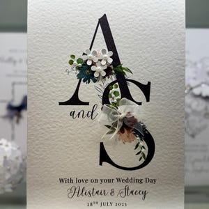 Pode incluir: Cartão de convite de casamento com as letras "A" e "S" em preto, adornadas com flores. O cartão inclui as palavras "and", "With love on your Wedding Day", e os nomes "Alistair & Stacey" com a data "28th July 2025".