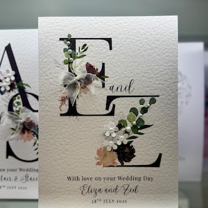 Puede incluir: Tarjeta de invitación de boda con las letras "E" y "Z" adornadas con flores y una cinta. La tarjeta dice "With love on your Wedding Day Eliza and Zed 28th July 2025". El fondo es de color blanco roto texturizado.