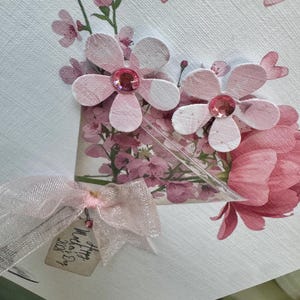 以下が含まれることがあります： ピンク色の紙の花、ラインストーンの中心、透明なリボンで作られた手作りの母の日カード。カードには「Happy Mother's Day 2026」と書かれたタグが付いています。デザインは花の要素とピンクのハートのアクセントが特徴です。