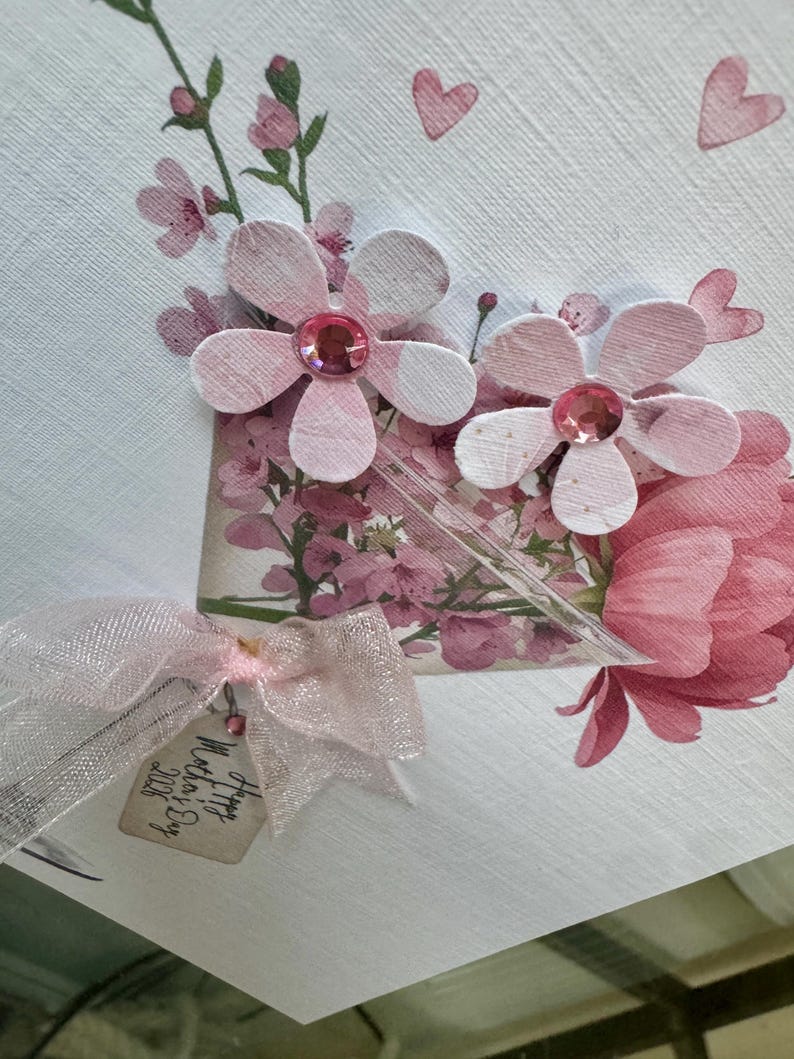 以下が含まれることがあります： ピンク色の紙の花、ラインストーンの中心、透明なリボンで作られた手作りの母の日カード。カードには「Happy Mother's Day 2026」と書かれたタグが付いています。デザインは花の要素とピンクのハートのアクセントが特徴です。