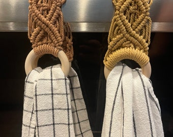 May include: Two macrame towel holders with wooden rings hanging on a stainless steel oven door. The towel holders are brown and mustard yellow. The towels are white with black stripes.