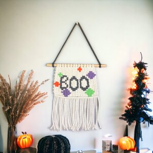 May include: A white macrame wall hanging with the word "BOO" in black yarn and colorful accents. The hanging is attached to a wooden dowel rod and has a fringe border.