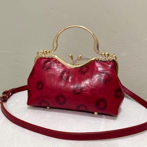 May include: A red leather handbag with a gold-colored clasp and handle. The bag features a floral pattern and a detachable shoulder strap. The handle is adorned with rhinestones, adding a touch of elegance to the design.
