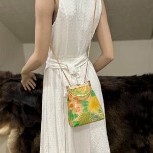 May include: A vintage handbag with a gold chain strap. The bag features a floral design in shades of yellow, green, and orange on a beige background. The bag has a gold-colored frame and clasp. The dress is white.
