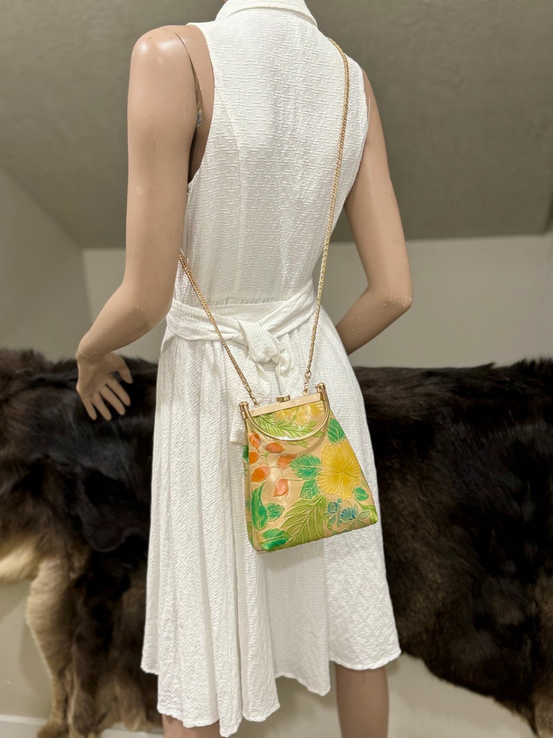 May include: A vintage handbag with a gold chain strap. The bag features a floral design in shades of yellow, green, and orange on a beige background. The bag has a gold-colored frame and clasp. The dress is white.
