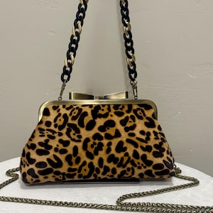May include: A structured handbag featuring a tan and black leopard print pattern with a textured finish. The bag has a brass-toned metal frame with a bow-shaped clasp, a detachable handle made of alternating black and gold links, and a long chain strap.