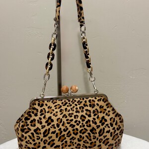 May include: A leopard print handbag with a kiss-lock closure. The bag features a chain strap with tortoise-shell-style links and a matching leopard print handle. The frame is silver-toned, and the clasp has two round, light-colored beads.