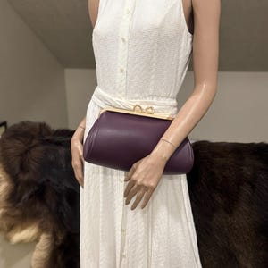 May include: A dark purple leather clutch bag with a gold-toned metal frame and a double-loop clasp closure. The bag features a pebbled texture and a curved, elongated shape, held against a white sleeveless collared dress with a textured fabric.