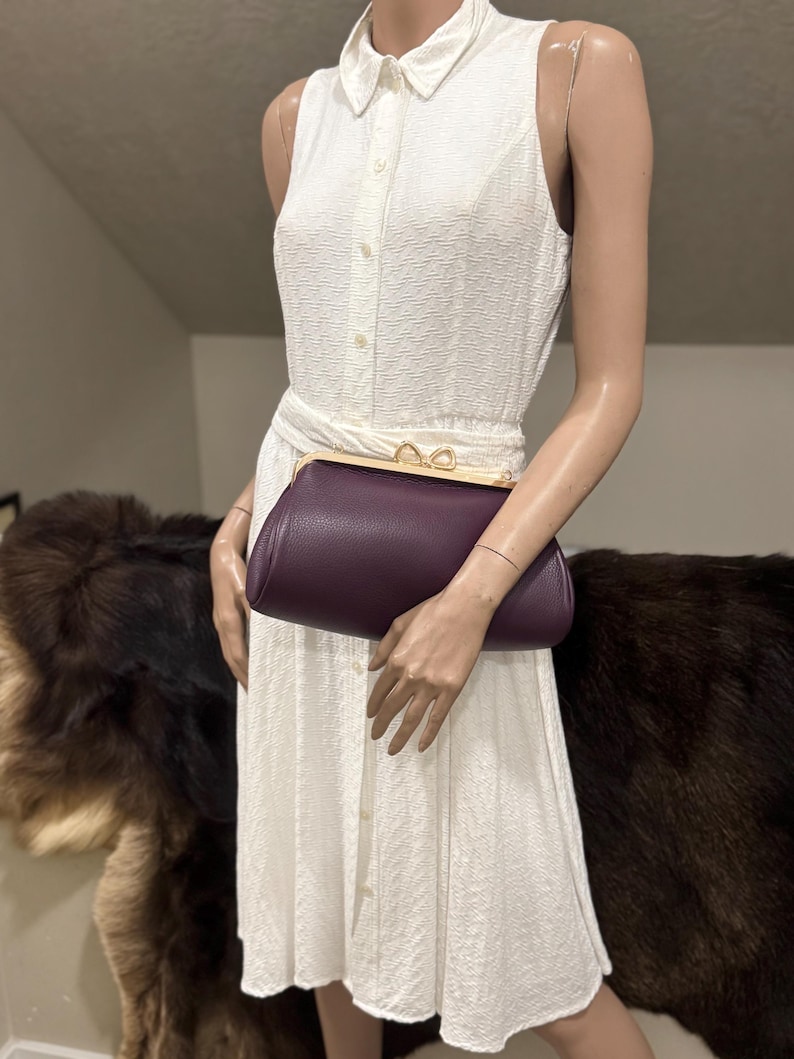 May include: A dark purple leather clutch bag with a gold-toned metal frame and a double-loop clasp closure. The bag features a pebbled texture and a curved, elongated shape, held against a white sleeveless collared dress with a textured fabric.