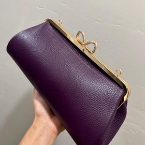 May include: A deep purple clutch bag with a textured surface and a gold-toned metal frame. The closure features a distinctive bow-shaped clasp at the top center, and there are small metal loops on the frame for attaching a strap to the accessory.