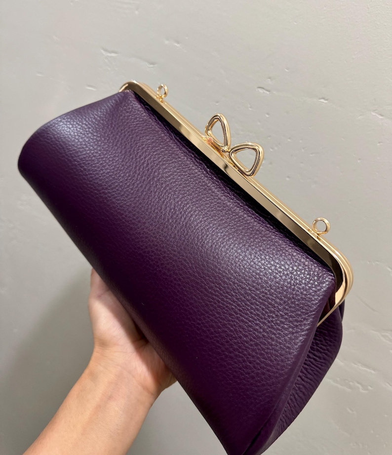 May include: A deep purple clutch bag with a textured surface and a gold-toned metal frame. The closure features a distinctive bow-shaped clasp at the top center, and there are small metal loops on the frame for attaching a strap to the accessory.