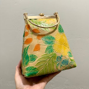 May include: Tan handbag with a floral design featuring yellow hibiscus flowers, green leaves, and orange accents. The bag has a gold-colored handle and clasp. The bag is held by a hand.