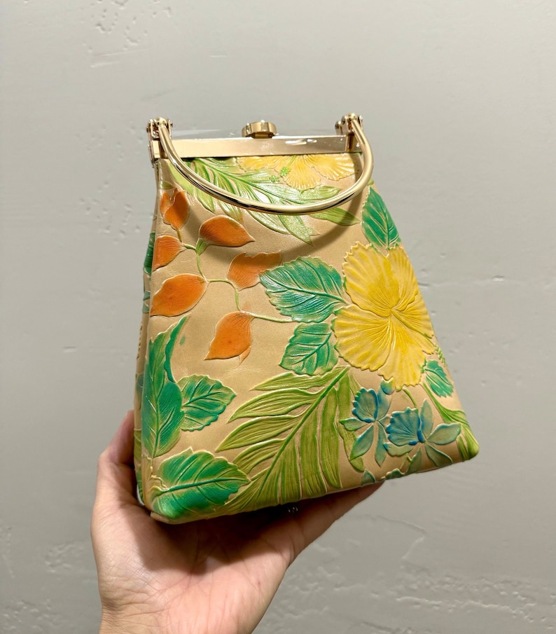 May include: Tan handbag with a floral design featuring yellow hibiscus flowers, green leaves, and orange accents. The bag has a gold-colored handle and clasp. The bag is held by a hand.