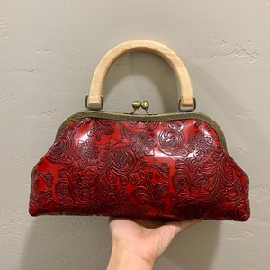 May include: A red, floral-patterned handbag with a wooden handle. The bag features a kiss-lock clasp and a detailed embossed design. The handle is a light-colored wood, and the metal accents are a bronze color.