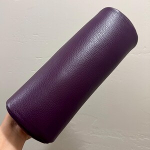 May include: A cylindrical bolster pillow featuring a deep purple exterior with a distinct pebbled texture. The item is elongated and rounded at both ends, showcasing a consistent solid color throughout its entire surface area.