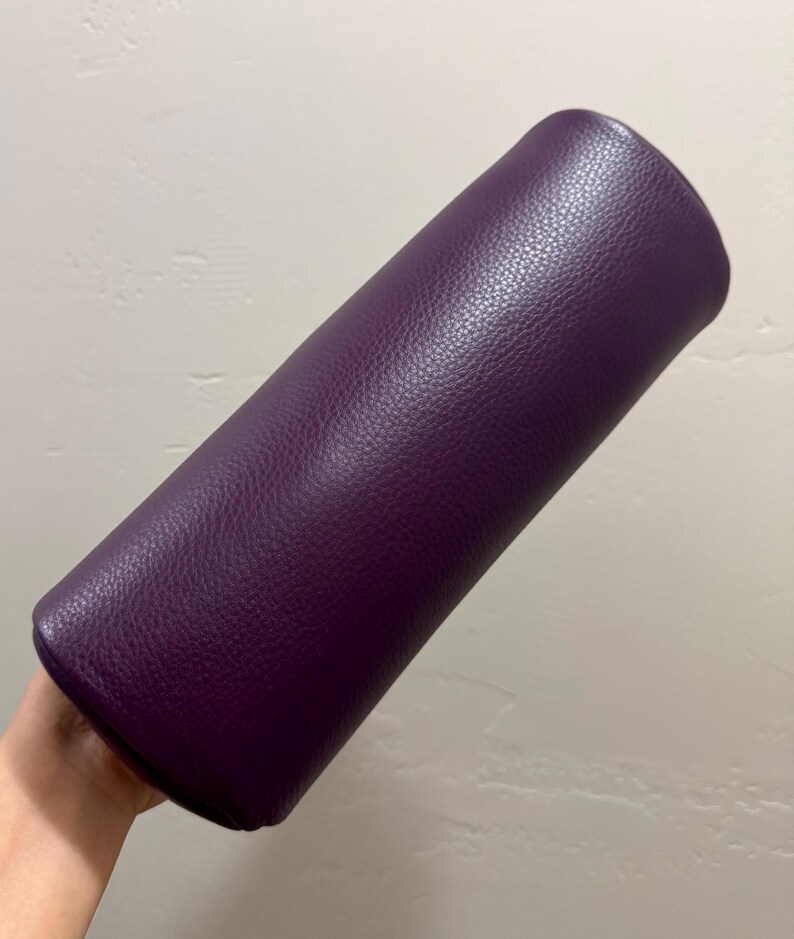 May include: A cylindrical bolster pillow featuring a deep purple exterior with a distinct pebbled texture. The item is elongated and rounded at both ends, showcasing a consistent solid color throughout its entire surface area.