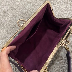 May include: A deep purple handbag with a gold-toned metal frame and a kiss-lock closure. The interior is lined with a matching purple fabric and features a single open pocket. The bag includes a gold-toned chain link strap and a purple handle.