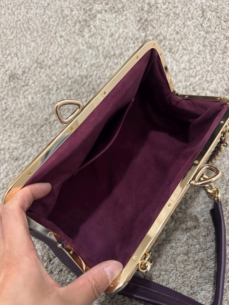 May include: A deep purple handbag with a gold-toned metal frame and a kiss-lock closure. The interior is lined with a matching purple fabric and features a single open pocket. The bag includes a gold-toned chain link strap and a purple handle.