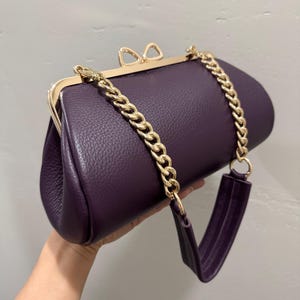 May include: A deep purple textured handbag with a gold-toned metal frame and a bow-shaped clasp. The bag features a gold-toned chain link shoulder strap that transitions into a purple leather-like material section for comfortable carrying.