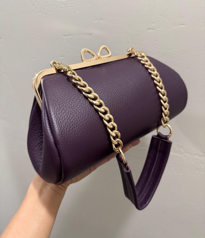 May include: A deep purple textured handbag with a gold-toned metal frame and a bow-shaped clasp. The bag features a gold-toned chain link shoulder strap that transitions into a purple leather-like material section for comfortable carrying.