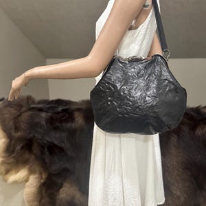 May include: A black handbag with a crinkled, textured surface and a silver-toned metal frame closure. The bag features a single shoulder strap and a smooth, oval-shaped metallic accent centered on the top clasp mechanism of the frame.
