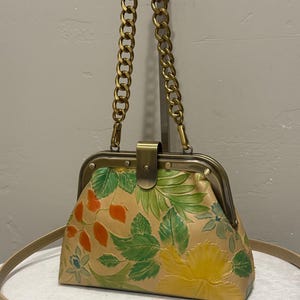 May include: A structured handbag featuring a tan base with embossed floral patterns in green, yellow, and orange. The bag has a brass-toned metal frame, a central clasp closure, and a thick gold-toned chain handle, complemented by a thin tan shoulder strap.