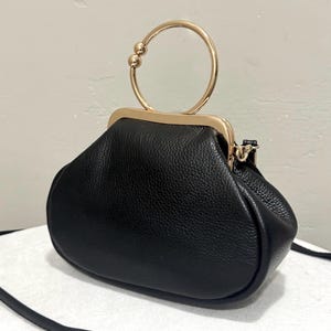 May include: Black handbag with a gold-colored handle. The bag has a rounded shape and a textured surface. The handle is a circular design with two small spheres at the top. A thin black strap is attached.