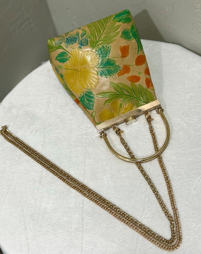 May include: A vintage handbag with a gold chain strap and a gold handle. The bag features a floral pattern with yellow hibiscus flowers, green leaves, and orange accents on a tan background. The bag has a rectangular shape.