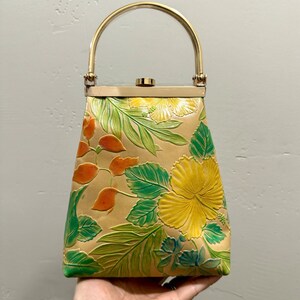 May include: Tan handbag with a gold handle and clasp. The bag features embossed floral designs in yellow, green, and orange. The bag is held by a hand.