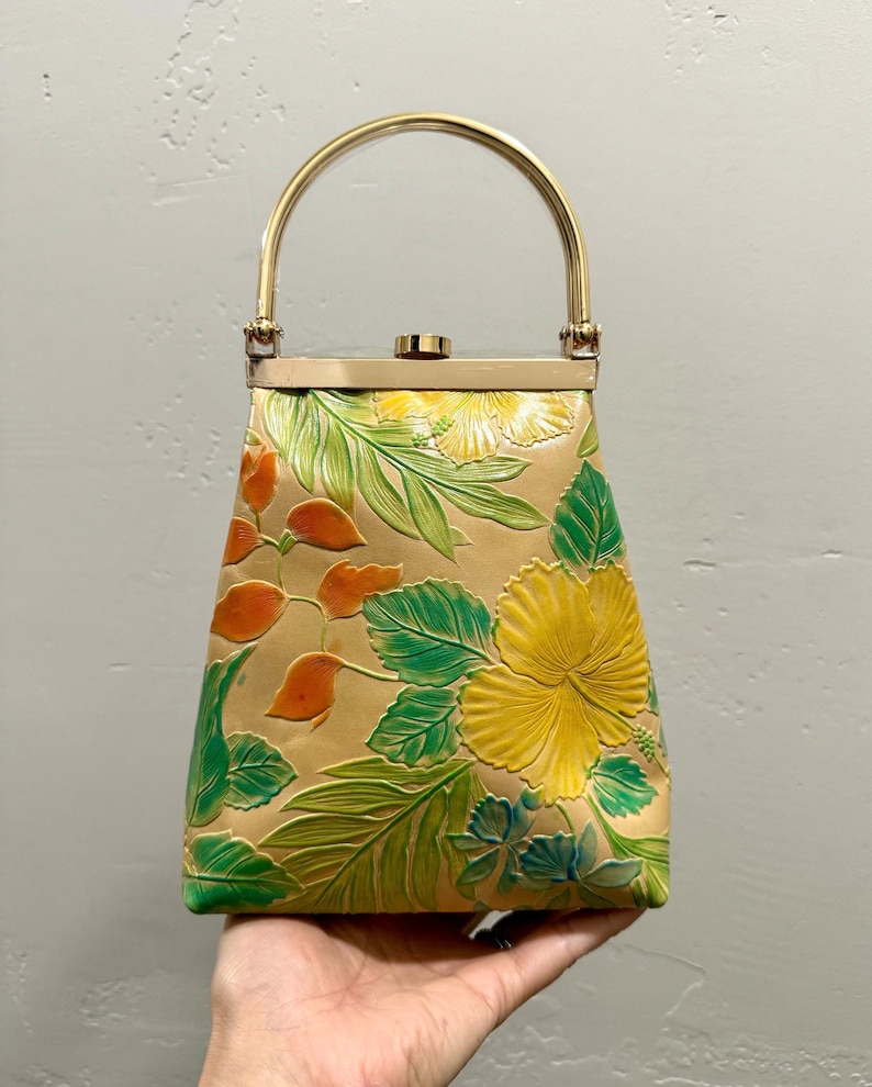 May include: Tan handbag with a gold handle and clasp. The bag features embossed floral designs in yellow, green, and orange. The bag is held by a hand.